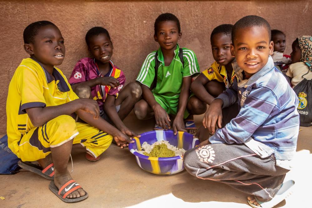 Children sit on the ground around a bowl of food and smile at the camera