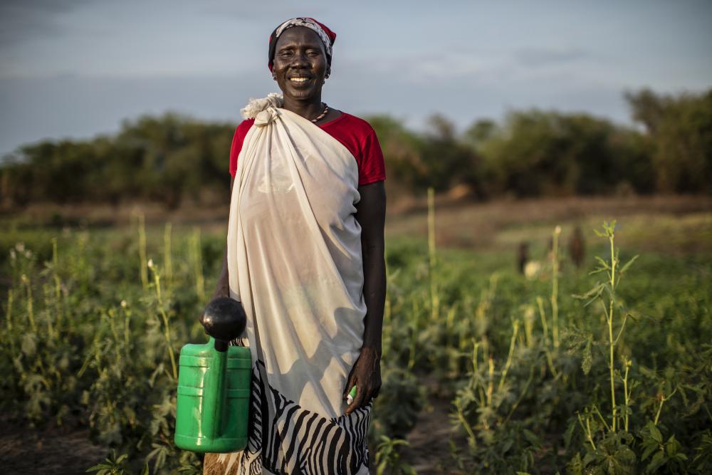 Woman from South Sudan stands in a field holding a watering can