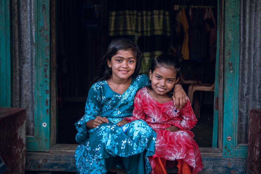 Two young Bangladeshi girls smile and sit together with their arms around each other
