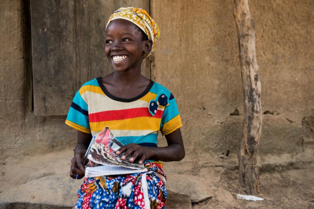 Niña sentada, sonriendo y sosteniendo un cuaderno y un lapicero. 